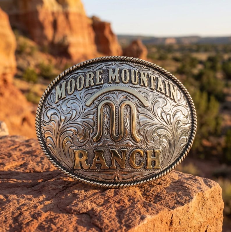 May include: An oval-shaped silver-tone belt buckle featuring an intricate engraved scrollwork pattern. The center displays a gold-tone letter M logo, with the text MOORE MOUNTAIN RANCH embossed in gold-tone lettering along the top and bottom edges.