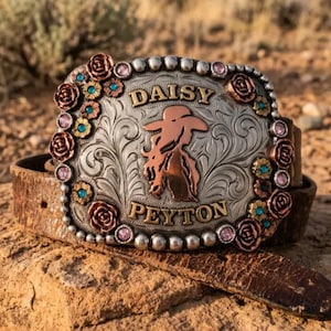 May include: A rectangular silver-toned belt buckle featuring an engraved floral pattern, copper-colored rose accents, and small turquoise and pink stones. The center displays a copper-colored silhouette of a person in a hat with the text DAISY PEYTON.