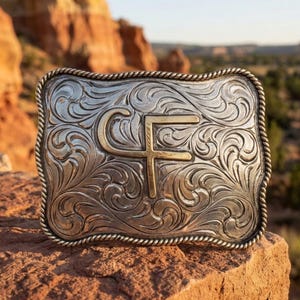 May include: A rectangular silver belt buckle with a rope-style border and intricate scrollwork engraving. The center features a prominent gold-toned letter C and F monogram. The surface has a polished finish with darkened recessed areas.