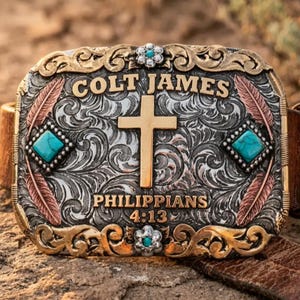 May include: A rectangular belt buckle featuring a silver-toned engraved scrollwork background with gold-toned accents. It displays the text COLT JAMES above a central gold cross and PHILIPPIANS 4:13 below. Two turquoise stones and two floral motifs are present.