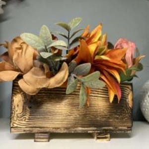 May include: A rustic wooden planter box filled with faux flowers in shades of orange, brown, and pink. The box has a distressed finish and sits on small wooden feet. Greenery accents the arrangement, creating a natural aesthetic.