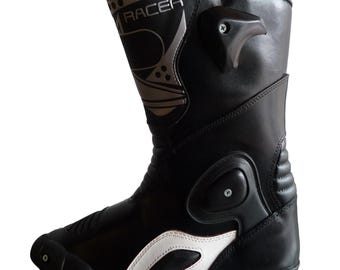 Könnte beinhalten: Schwarzer und weißer Motorradstiefel mit dem Wort "RACER" in Silber. Der Stiefel hat ein hoch geschnittenes Design mit verstärkten Einsätzen und einem weißen Akzent an der Seite. Der Stiefel ist aus Leder und hat eine Gummisohle.