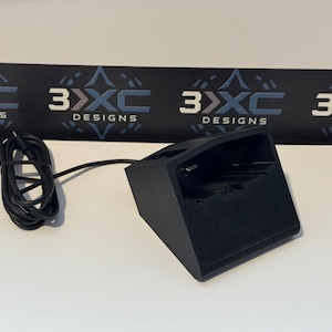 May include: A black charging dock with an angled front surface and a built-in USB-C connector. A black charging cable is attached to the side of the dock. The background features a black banner with the repeating white and blue text 3XC DESIGNS.
