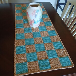 May include: A decorative table runner with a checkerboard pattern in teal and brown, topped with a white vase with colorful abstract paint strokes. The runner is placed on a dark brown wooden table.