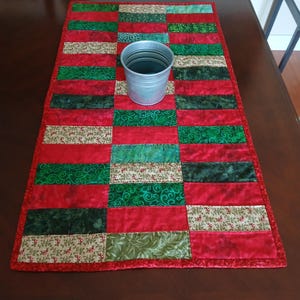 May include: A rectangular quilted table runner with a patchwork design. The runner features alternating red and green fabric blocks, some with floral patterns. A small, silver metal bucket sits on the runner. The runner is on a dark brown wooden table.