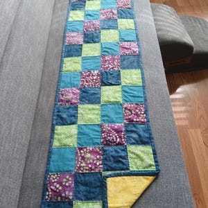 May include: A handmade, quilted table runner with a patchwork design. The runner features squares of teal, green, purple, and navy blue fabrics. The underside is yellow. The runner is approximately 60 inches long.