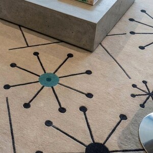 May include: Beige rug with a mid-century modern design featuring black starburst patterns and teal accents. A gray concrete coffee table with a book on top is in the background. The book's title is "anti Suga Senige".