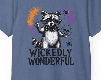 Op de afbeelding: Blauw t-shirt met een cartoon wasbeer die een schedel vasthoudt en een duim omhoog geeft. De wasbeer is omgeven door oranje en paarse accenten, met de tekst "WICKEDLY WONDERFUL" eronder.