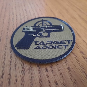May include: Circular patch with a black pistol silhouette and a target reticle above it. The text "TARGET ADDICT" is below the gun. The patch has a dark blue border and a tan background.