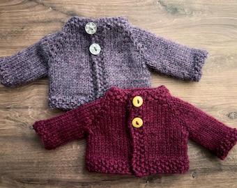 May include: Two small, long-sleeved knit cardigans. The top cardigan is a heathered purple with two clear, round buttons. The bottom cardigan is a solid deep burgundy with two yellow, round buttons. Both garments feature a ribbed texture.