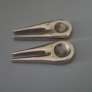 以下が含まれることがあります： 2つの銀色のゴルフディボット修理ツール。各ツールは、尖った端と円形のくぼみがあります。ツールは灰色の表面に配置されており、ゴルフアクセサリーや機器に焦点を当てていることを示唆しています。