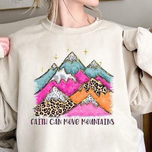 Könnte beinhalten: Beiges Sweatshirt mit einem farbenfrohen Berg-Motiv. Das Design zeigt türkisfarbene, rosa, orangefarbene Berge und Berge mit Leopardenmuster mit goldenen Akzenten und dem Text "Faith Can Move Mountains" in dunkler violetter Schrift.