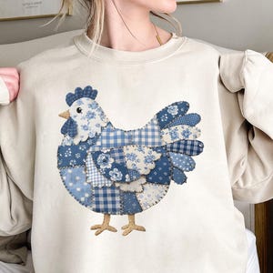 May include: Cream-coloured sweatshirt with a patchwork chicken design in various shades of blue. The chicken features floral patterns, gingham, and solid blue patches. The sweatshirt has a crew neck and long sleeves.
