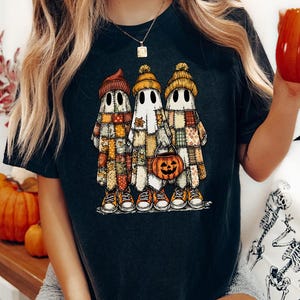 Könnte beinhalten: Ein schwarzes T-Shirt mit einer Grafik von drei Geistern, die bunte, im Patchwork-Stil gemusterte Gewänder und Mützen tragen. Die Geister tragen Turnschuhe und der mittlere Geist hält einen kleinen orangefarbenen Kürbis-Eimer.