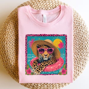 May include: A light pink t-shirt featuring a graphic of a leopard wearing sunglasses and a sun hat, floating in a pink inner tube. The graphic is set against a turquoise background with palm trees and a yellow sun. The shirt is on a woven surface.