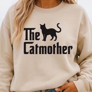 May include: Beige sweatshirt with the text "The Catmother" and a black cat silhouette. The text is in a bold, stylized font. The sweatshirt is a neutral color, and the overall design is simple and graphic.