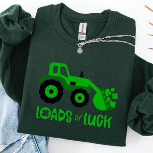 Może przedstawiać: Ciemnozielona bluza z nadrukiem zielonego traktora i napisem "Loads of Luck" w kolorze zielonym. Traktor ma czarne koła i łyżkę wypełnioną zielonymi koniczynami. Srebrny naszyjnik jest zarzucony na bluzę.