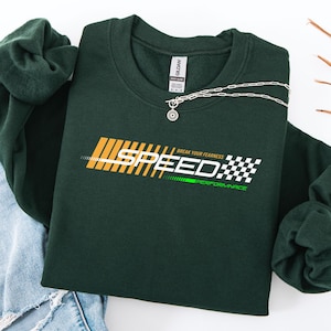 May include: A dark green sweatshirt with the word "SPEED" in yellow and white block letters. The text "BREAK YOUR FEARNESS PERFORMANCE" is below the word "SPEED". A silver necklace with a pendant is on the sweatshirt.