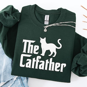 May include: A dark green sweatshirt with the text "The Catfather" and a white cat silhouette. A silver necklace with a pendant is draped across the sweatshirt. The sweatshirt is paired with blue jeans.