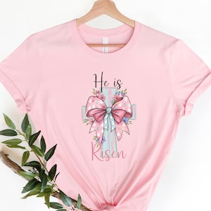 Puede incluir: Camiseta rosa claro con un diseño de cruz, un lazo rosa y detalles florales. Las palabras "He is Risen" están impresas en negro. La camiseta está colgada en una percha de madera.