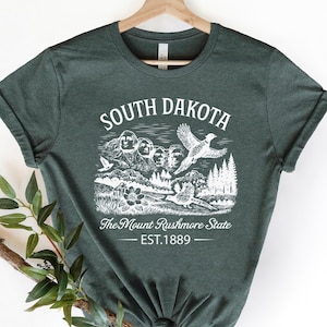 May include: Dark green t-shirt with a white graphic featuring the text "SOUTH DAKOTA" and an illustration of Mount Rushmore, a flying bird, and trees. The text "The Mount Rushmore State" and "EST. 1889" are also included in the design.