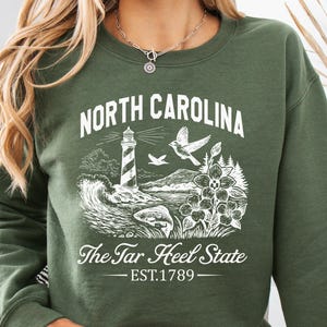 Op de afbeelding: Groen sweatshirt met een witte afbeelding van een vuurtoren, golven, vogels en bloemen. De tekst "North Carolina" en "The Tar Heel State" zijn boven en onder de afbeelding gedrukt, met "EST.1789" eronder.