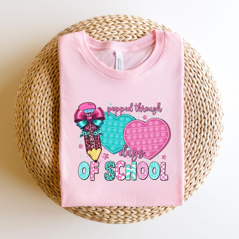 May include: A light pink t-shirt with the text "popped through days of school". The shirt features a pencil with a bow, and two heart-shaped pop-it toys in teal and pink. The shirt is folded on a woven surface.