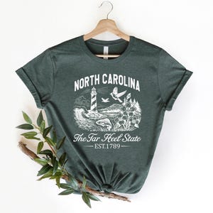 Op de afbeelding: Donkergroen t-shirt met een witte afbeelding van de kustlijn van North Carolina, inclusief een vuurtoren, vogels en bloemen. De tekst "NORTH CAROLINA" en "The Tar Heel State" staan boven en onder de afbeelding, met "EST.1789" eronder.
