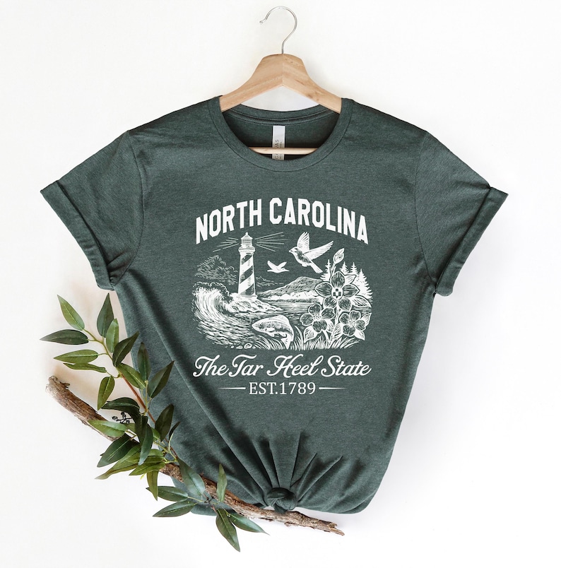 Op de afbeelding: Donkergroen t-shirt met een witte afbeelding van de kustlijn van North Carolina, inclusief een vuurtoren, vogels en bloemen. De tekst "NORTH CAROLINA" en "The Tar Heel State" staan boven en onder de afbeelding, met "EST.1789" eronder.
