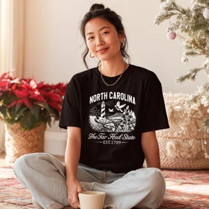 Op de afbeelding: Zwart t-shirt met een witte afbeelding van een vuurtoren, golven en de tekst "North Carolina The Tar Heel State EST.1789". De persoon draagt het t-shirt en zit op een tapijt.
