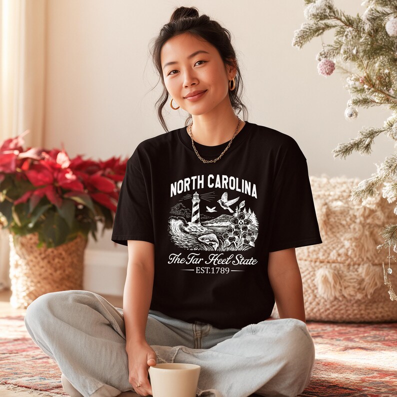 Op de afbeelding: Zwart t-shirt met een witte afbeelding van een vuurtoren, golven en de tekst "North Carolina The Tar Heel State EST.1789". De persoon draagt het t-shirt en zit op een tapijt.