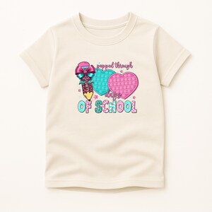 May include: Cream-colored t-shirt featuring a colorful graphic. The design includes a pencil with a bow, two heart-shaped pop-it toys, and the text "popped through days OF SCHOOL". The graphic has pink, blue, and teal accents.