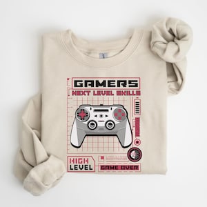 Könnte beinhalten: Beiges Sweatshirt mit einem Grafikdesign, das einen Videospiel-Controller zeigt. Das Design enthält die Wörter "GAMERS", "NEXT LEVEL SKILLS", "HIGH LEVEL" und "GAME OVER" in Rot und Schwarz. Der Controller ist grau und weiß.