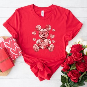 Könnte beinhalten: Ein rotes T-Shirt mit einer Cartoonfigur, die eine herzförmige Sonnenbrille trägt und ein Herz mit dem Wort "LOVE" hält. Das Design umfasst Herzen und ein Patchworkmuster. Das T-Shirt ist an der Taille geknotet.