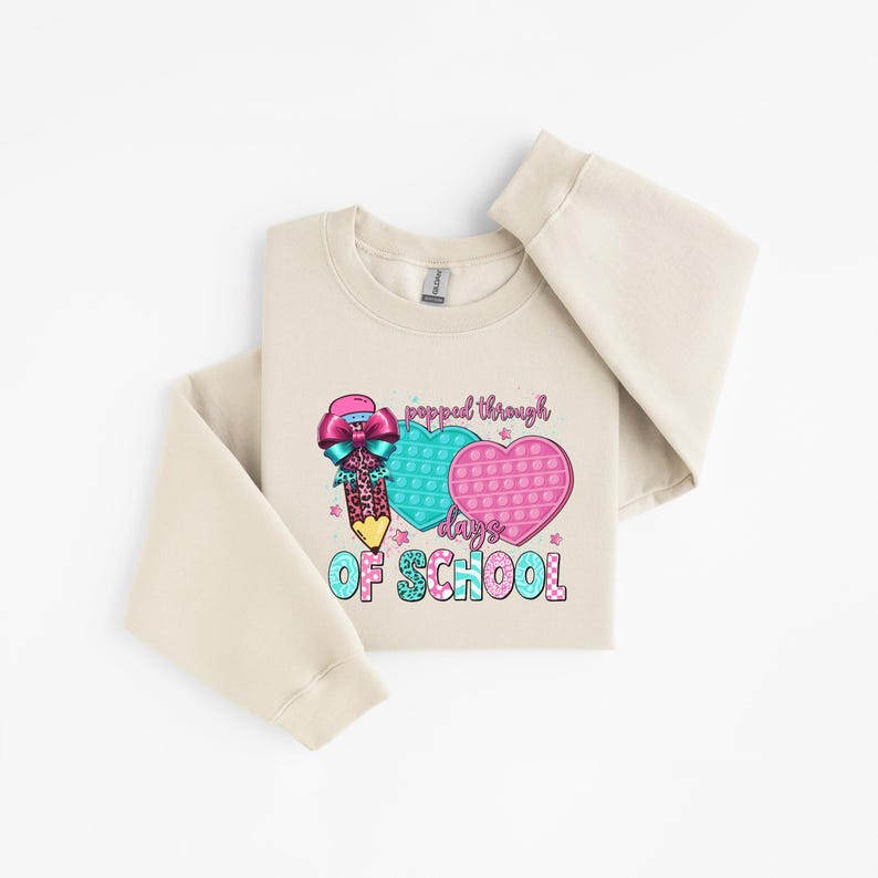 May include: A cream-colored sweatshirt with a colorful graphic. The graphic features a pencil with a bow, two heart-shaped pop-it toys, and the text "popped through days of school". The design includes shades of pink, teal, and purple.