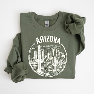 Può includere: Felpa verde oliva con grafica bianca di un paesaggio desertico dell'Arizona, con cactus e montagne, all'interno di una cornice circolare. La parola "ARIZONA" è arcuata sopra.