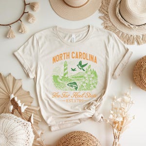 Op de afbeelding: Cr&egrave;mekleurig T-shirt met een oranje en groen grafisch ontwerp. Het ontwerp bevat de woorden "North Carolina", een vuurtoren, vogels en de tekst "The Tar Heel State". Het oprichtingsjaar "EST.1789" is ook aanwezig.