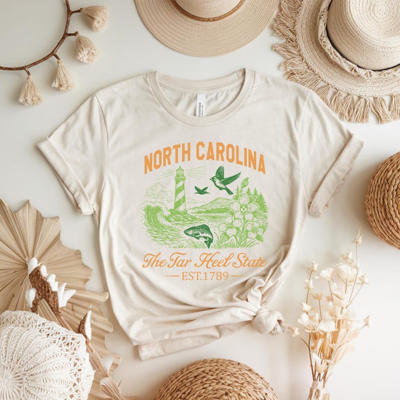 Op de afbeelding: Cr&egrave;mekleurig T-shirt met een oranje en groen grafisch ontwerp. Het ontwerp bevat de woorden "North Carolina", een vuurtoren, vogels en de tekst "The Tar Heel State". Het oprichtingsjaar "EST.1789" is ook aanwezig.