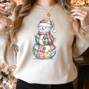 May include: A cream-colored sweatshirt featuring a whimsical snowman design. The snowman is adorned with a patchwork body, a colorful hat, and a striped scarf. The design includes button details and string lights, perfect for the holiday season.