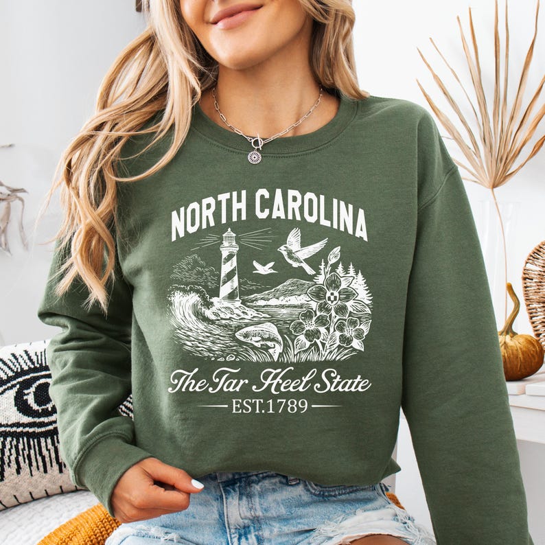 Op de afbeelding: Groen sweatshirt met een witte afbeelding van een vuurtoren, golven, vogels en bloemen. De tekst "North Carolina" en "The Tar Heel State" zijn boven en onder de afbeelding gedrukt, met "EST.1789" eronder.