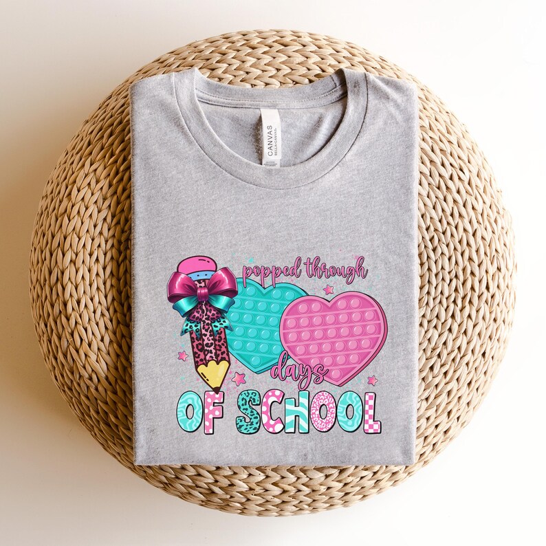 May include: A gray t-shirt with a colorful graphic design. The design includes a pencil, two heart-shaped pop-it toys, and the text "popped through 100 days of school". The shirt is displayed on a woven, circular surface.