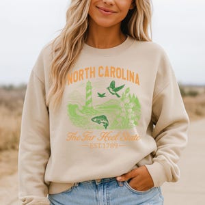 Op de afbeelding: Beige sweatshirt met een North Carolina-ontwerp. De afbeelding bevat een vuurtoren, vogels en vissen in groen en oranje. Tekst luidt "North Carolina" en "The Tar Heel State EST.1789". Een relaxte, comfortabele top.