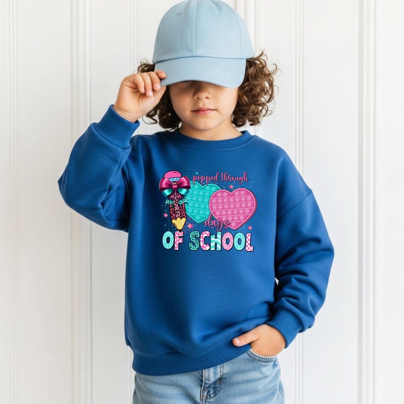 May include: A blue sweatshirt with a graphic that says "popped through days of school". The graphic includes two heart-shaped pop-it toys, one teal and one pink, and a pencil with a bow. The child is wearing a light blue baseball cap.