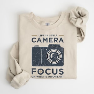 May include: Beige sweatshirt with the text "Life is like a Camera, Focus on What's Important" and a navy blue camera graphic. The sweatshirt has long sleeves and a crew neck. The design is printed on the front.