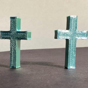 Może przedstawiać: Dwa turkusowe krzyże są wyświetlane na neutralnym tle. Krzyże mają teksturowaną powierzchnię i prosty, tradycyjny design. To prawdopodobnie symbole religijne.