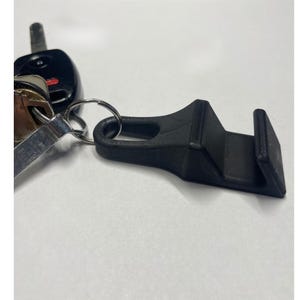 Puede incluir: Un clip negro impreso en 3D para el cinturón de seguridad del coche, sujeto a un llavero con llaves del coche. El clip tiene un diseño curvo con una ranura para el cinturón de seguridad y un lazo para el llavero.