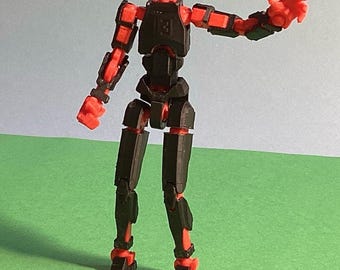 Op de afbeelding: Een zwart en rood gearticuleerd actiefiguur met het label DUMMY 13 3D PRINTED ACTION FIGURE. Het figuur heeft een blokvormig hoofd, een borstplaat en gewrichtsleden met zichtbare rode verbindingen tussen de zwarte segmenten in een pose.