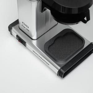 May include: A silver and black Moccamaster Cup-One coffee maker. The machine features a rectangular base with black trim, a black drip tray, and a silver water reservoir. The power switch is red. The text "Cup-one" is visible.