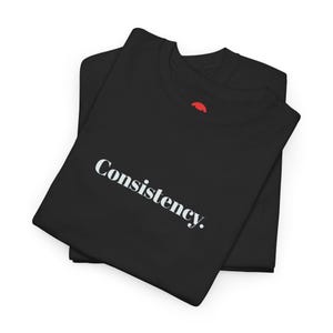 Könnte beinhalten: Zwei schwarze T-Shirts sind ordentlich gefaltet und gestapelt. Das obere Shirt zeigt das Wort „Consistency“ in weißer, serifenloser Schrift. Die Shirts sind aus einem weichen Material und werden auf weißem Hintergrund präsentiert.