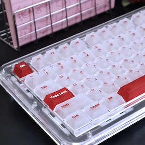 Puede incluir: Un teclado mecánico transparente de acrílico con teclas blancas y teclas de acento rojas. Las teclas tienen letras rojas. El teclado tiene una base transparente.